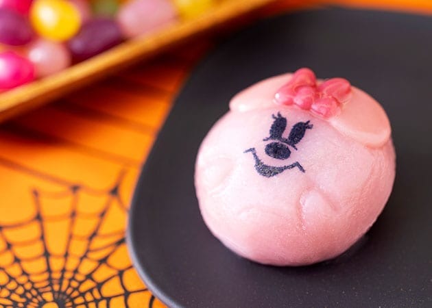 A pink, round dessert with a cartoon face and bow on a black plate—part of 2025 Halloween Merchandise at Tokyo Disney Resort.