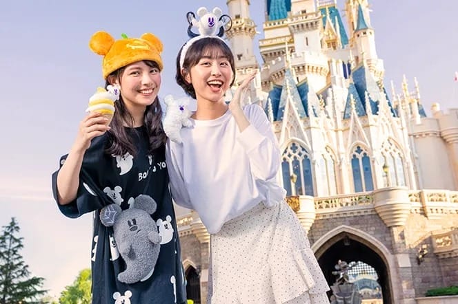 Two smiling women in character hats pose at Tokyo Disney Resort in front of a castle on a sunny day.