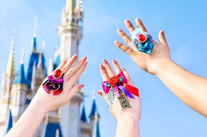 Three hands with colorful ribbon rings raised before Tokyo Disney Resort’s fairytale castle against a bright blue sky.