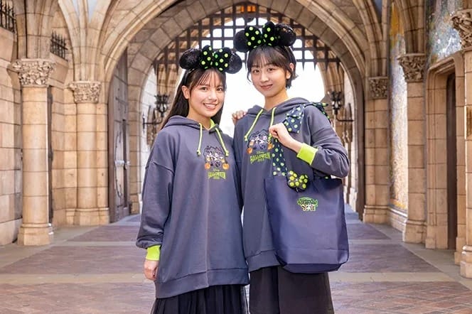 Two women in matching Minnie Mouse ear headbands and sweatshirts smile at Tokyo Disney Resort’s stone archway.