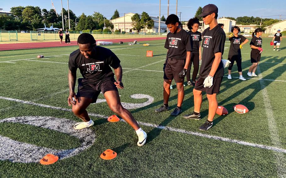 A student-athlete in a JFA American Football shirt demonstrates footwork drills through orange cones on a football field while several other athletes in matching black JFA shirts watch during a daytime practice session.