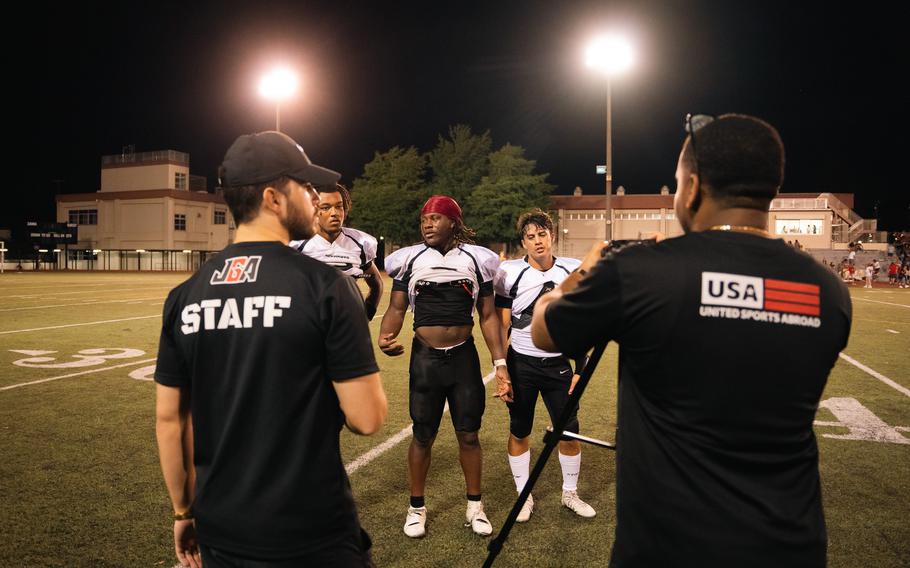 Two men in black shirts talk with three young football players in white and black uniforms on an illuminated football field at night, with residential buildings visible in the background.