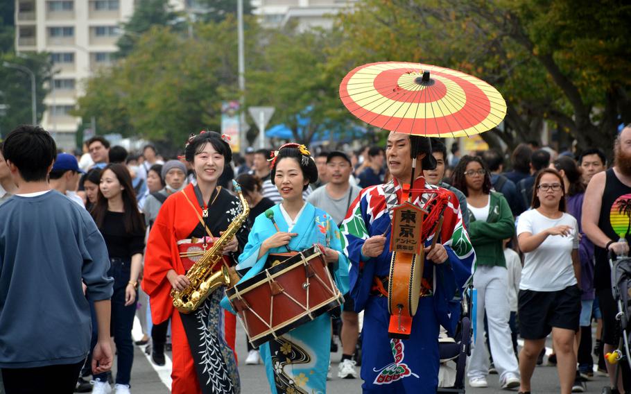 Three musicians wearing traditional Japanese clothing walk through a crowded street festival. The woman on the left wears a red and black yukata while holding a saxophone, the center performer wears a light blue kimono and carries a traditional drum, and the man on the right wears a colorful patterned kimono with a red and yellow paper umbrella balanced on his head.