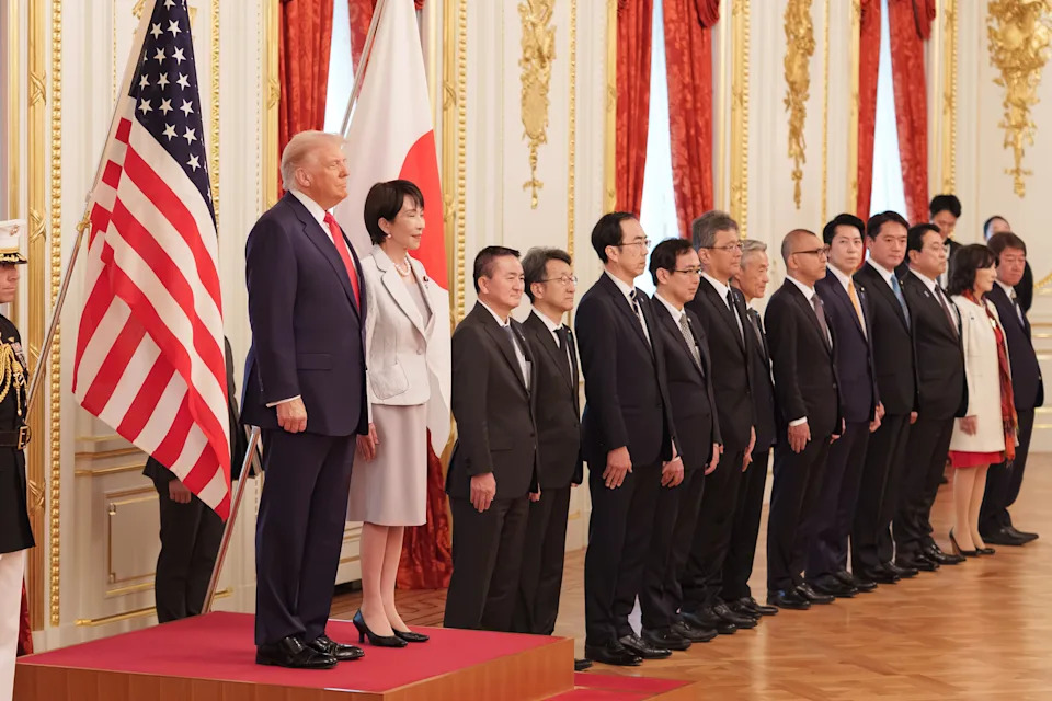 A group of officials, including a man and a woman on a platform, stand in a formal setting with U.S. and Japanese flags visible