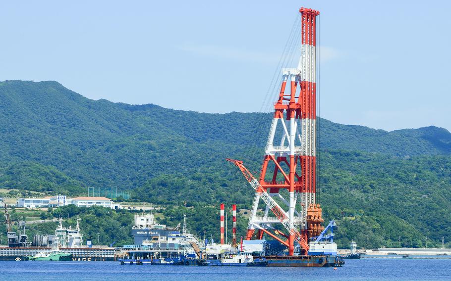 A large red and white construction crane extends upward from a vessel idling in a bay, with other construction ships around it and tree-covered hills in the background.