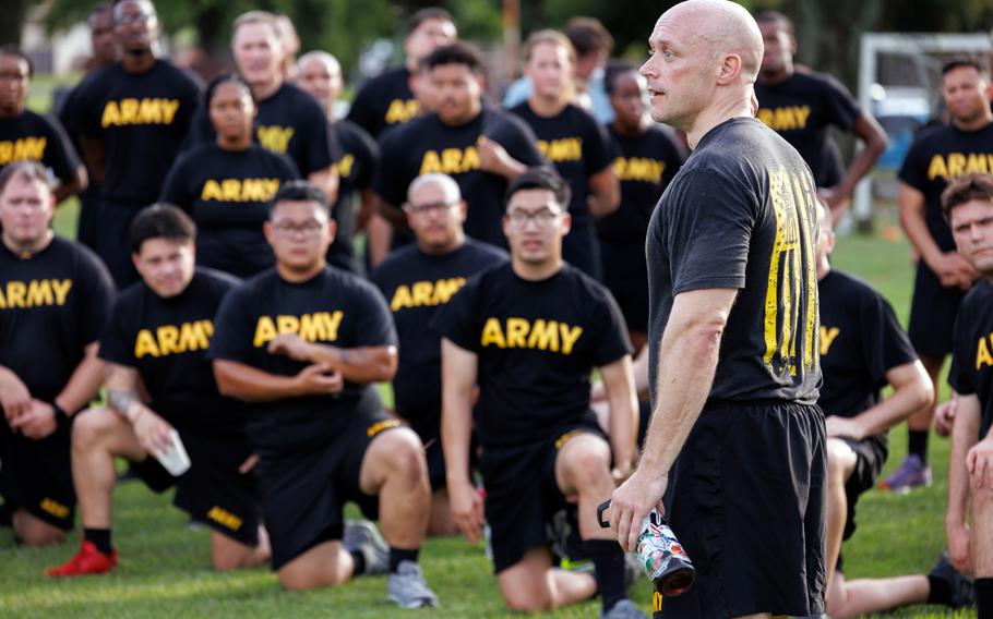 A man in an Army PT uniform (black T-shirt and shorts) stands addressing a large group of soldiers seated on grass. 