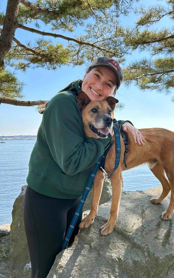 A woman smiles with a dog while standing in front of a body of water.