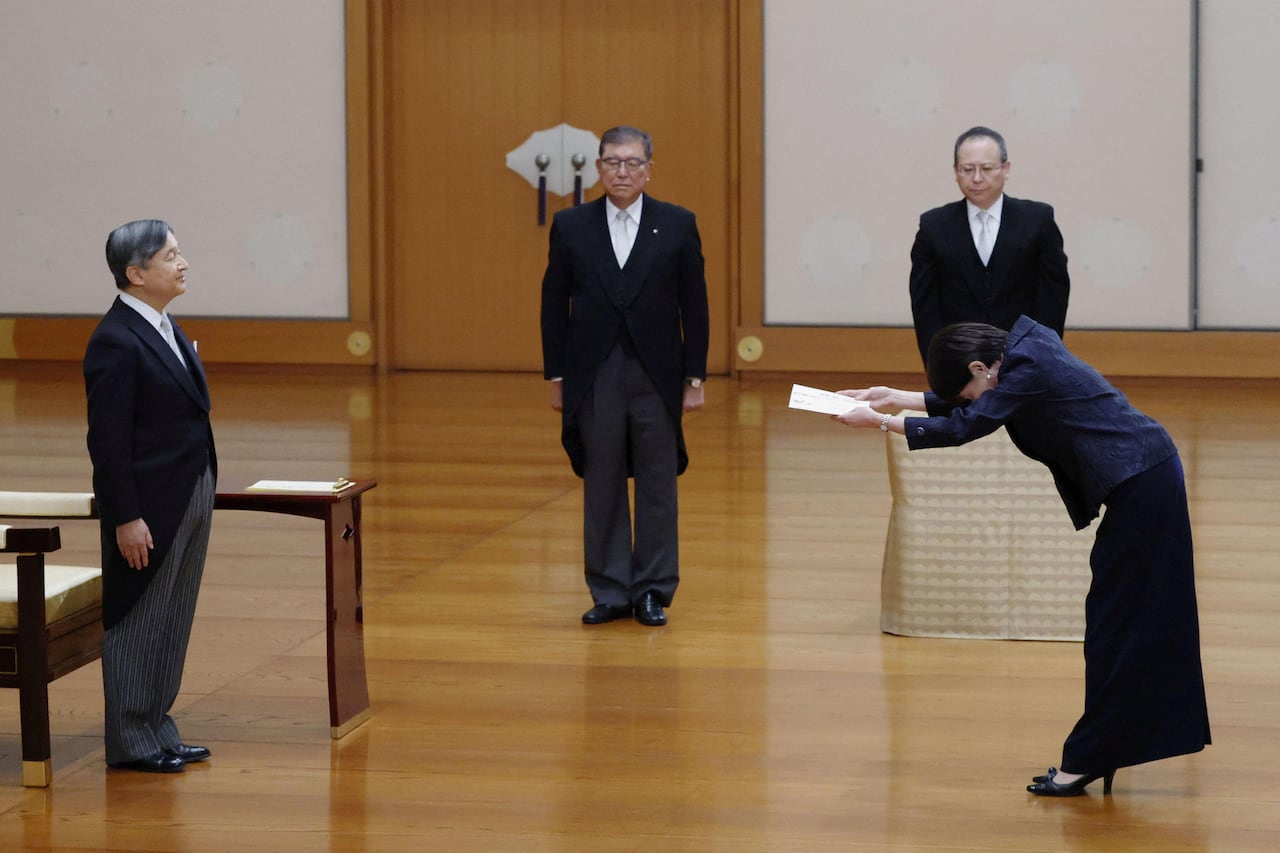 An Asian woman bows toward an older Asian man inside a room while holding a piece of paper. Two other Asian men in suits look on.