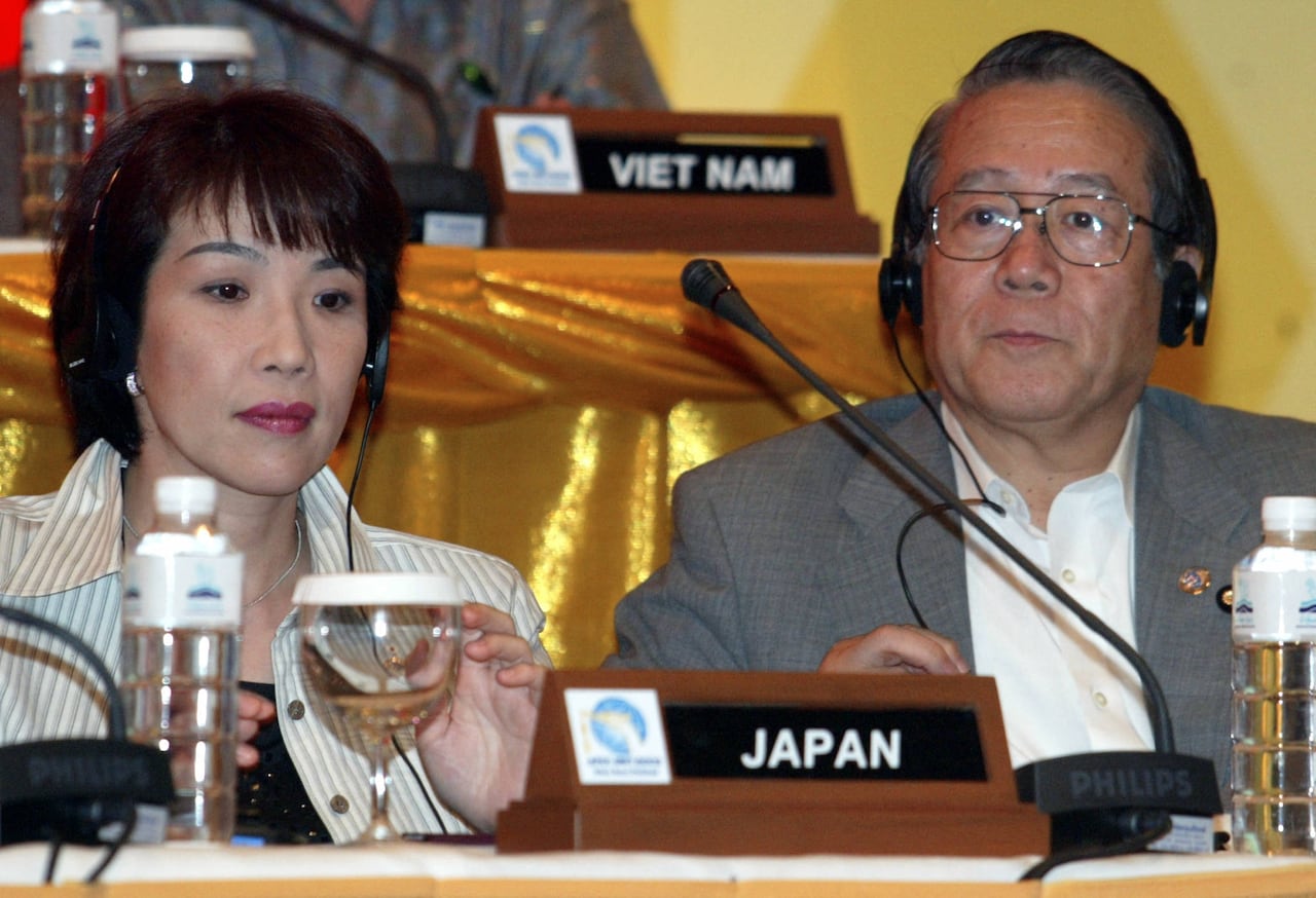 An Asian woman and an Asian man in glasses are shown seated behind a placard that says 'Japan.'