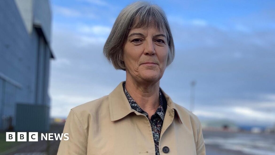 Julia Longbottom, a lady with short grey hair stares at the camera with a serious  expression. She is wearing a patterned blouse and a beige rain jacket. An industrial complex can be seen in the background.
