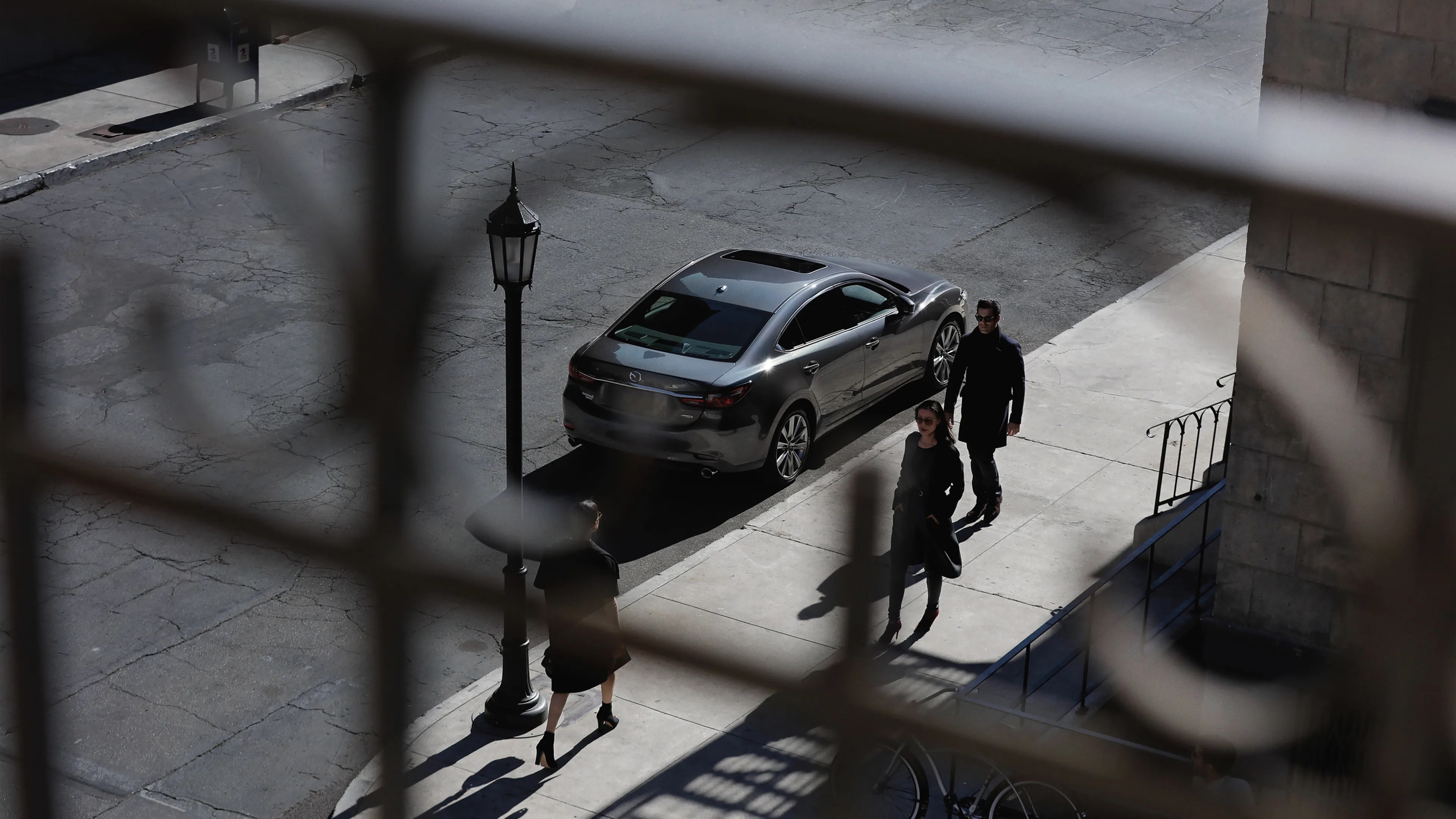 Dark gray sedan parked on a street next to a sidewalk with three people walking nearby, viewed through a metal fence.