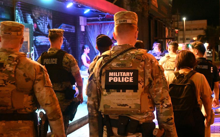 Military police officers in camouflage uniforms standing outside an establishment with colorful blue and red lighting at night. 