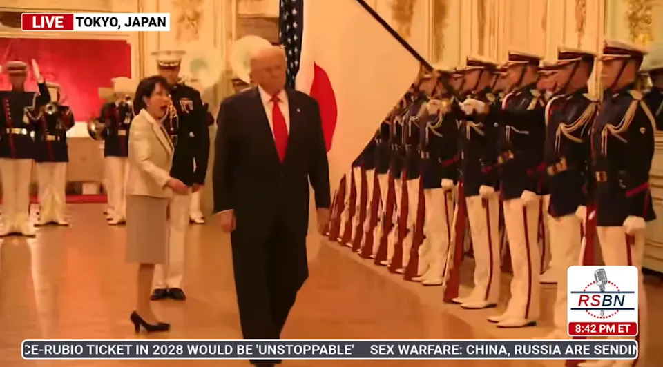 A group of officials and military personnel greet a man in a suit walking in a formal setting, captured during a broadcast in Tokyo, Japan