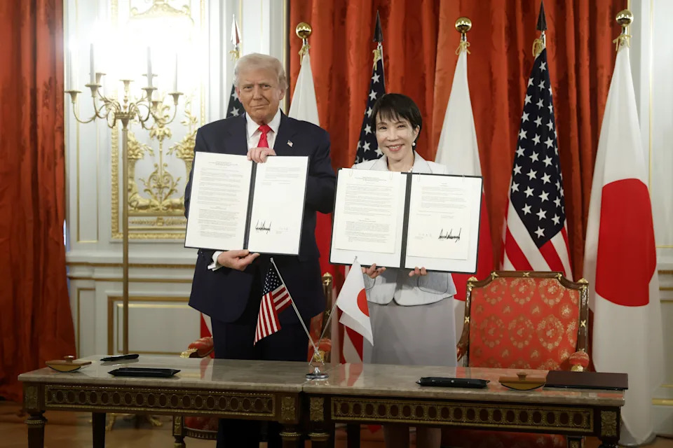 Two people stand holding signed documents in a formal setting with U.S. and Japanese flags in the background