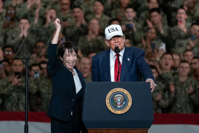 Two individuals at a podium, with one raising their arm. The person on the right is wearing a suit and a 'USA' cap. Audience in the background