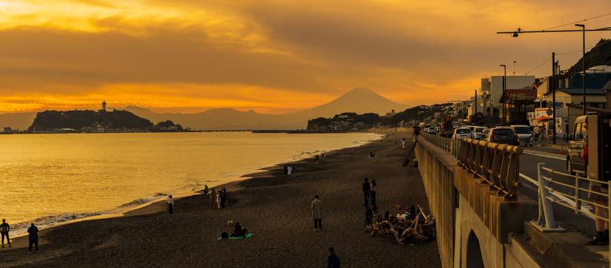 Golden sunset over Shonan Coast with silhouettes of Enoshima Island and Mount Fuji, beachgoers relaxing along the shore.