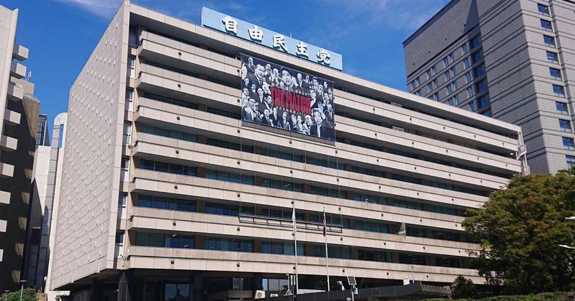 A large modern office building with a Japanese sign on the roof and a black-and-white banner featuring characters from the anime "Attack on Titan" displayed across its facade. Blue sky and other tall buildings are in the background.