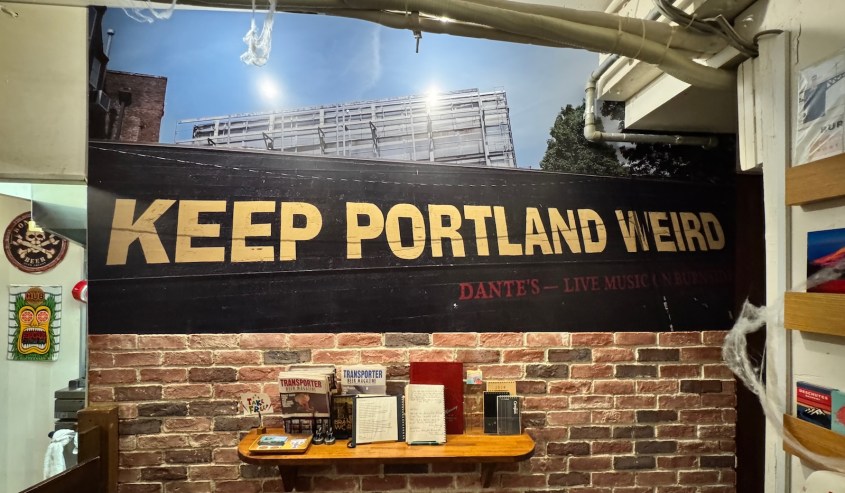 Wall mural displaying the phrase 'KEEP PORTLAND WEIRD' in large letters above a brick wall, with various memorabilia on a wooden table below.