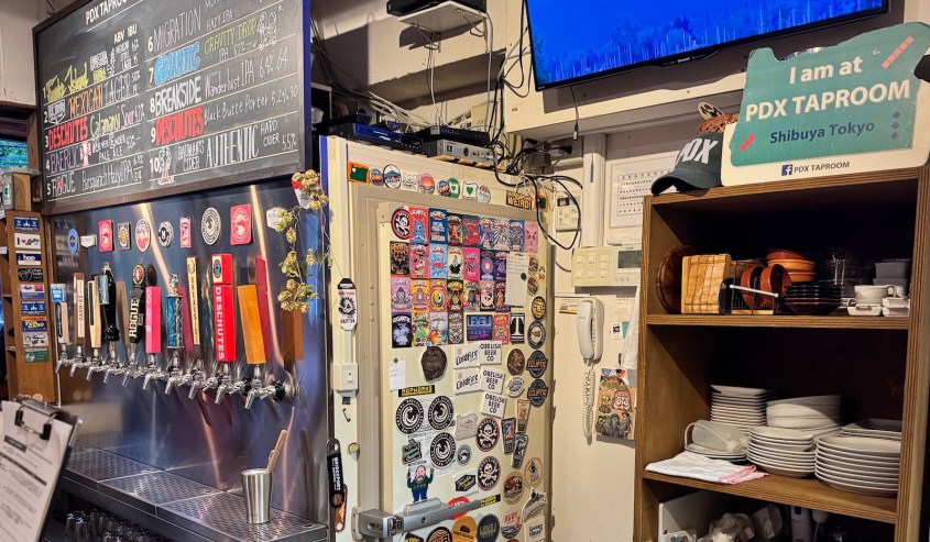 Interior of PDX Taproom in Shibuya, Tokyo, featuring a bar with craft beer taps, a wall covered in stickers, and a sign indicating the location.