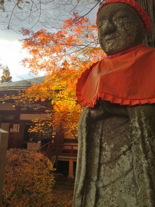 Stone statue wearing cloth bib and cap at Chusonji, Hiraizumi, with red autumn leaves in the background