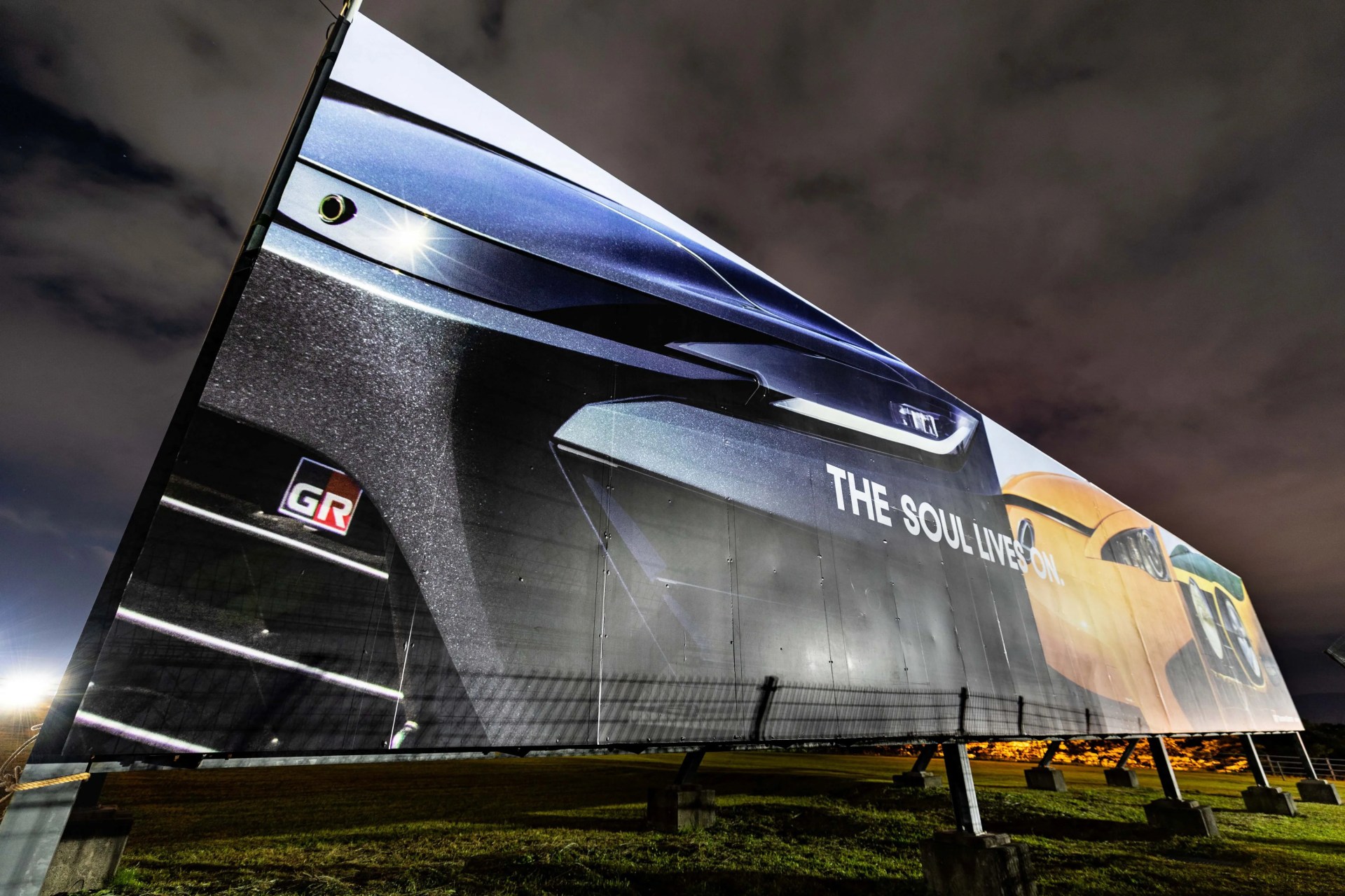 Large outdoor billboard at night showing close-up of a black car front with 