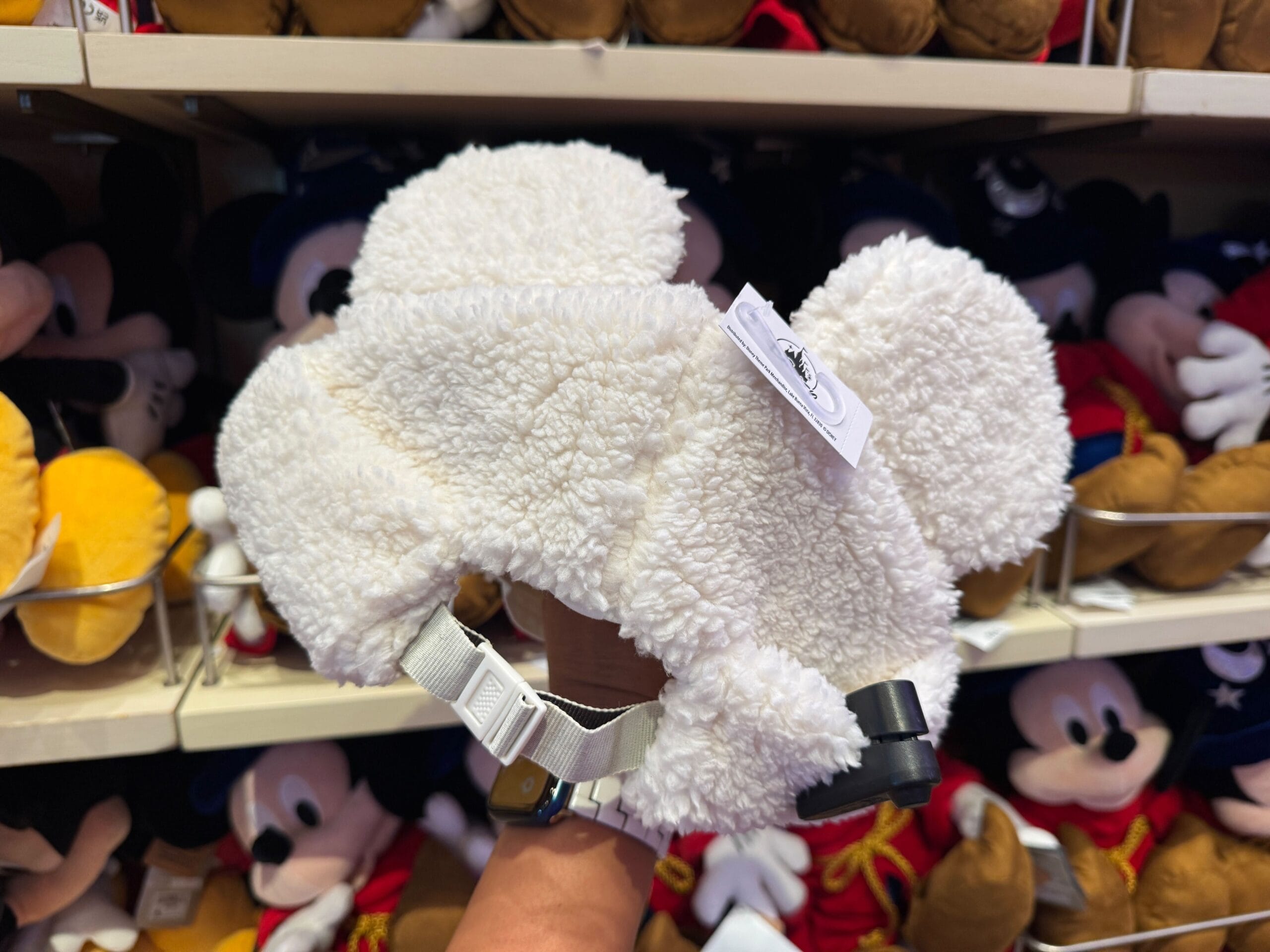 A hand holds a fluffy white Winter Cap with Mickey Mouse ears among Mickey plush toys in a store display.