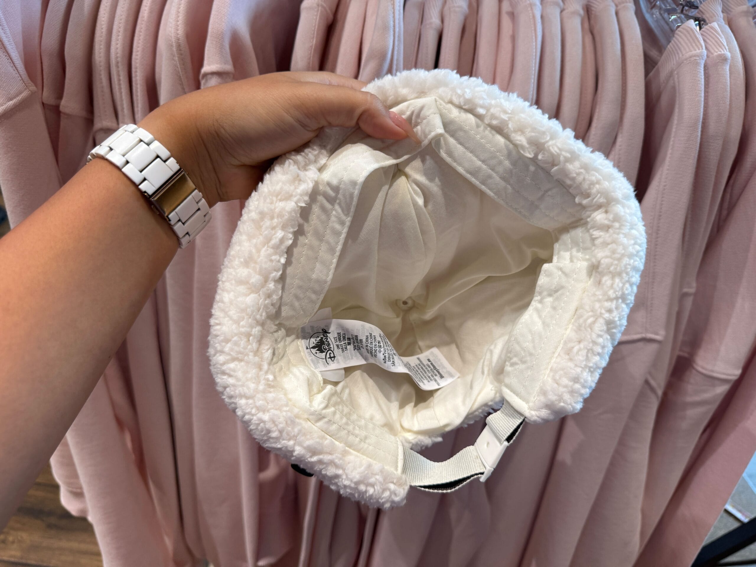 A hand holds a white, fluffy Winter Cap above pink shirts hanging on a clothing rack.