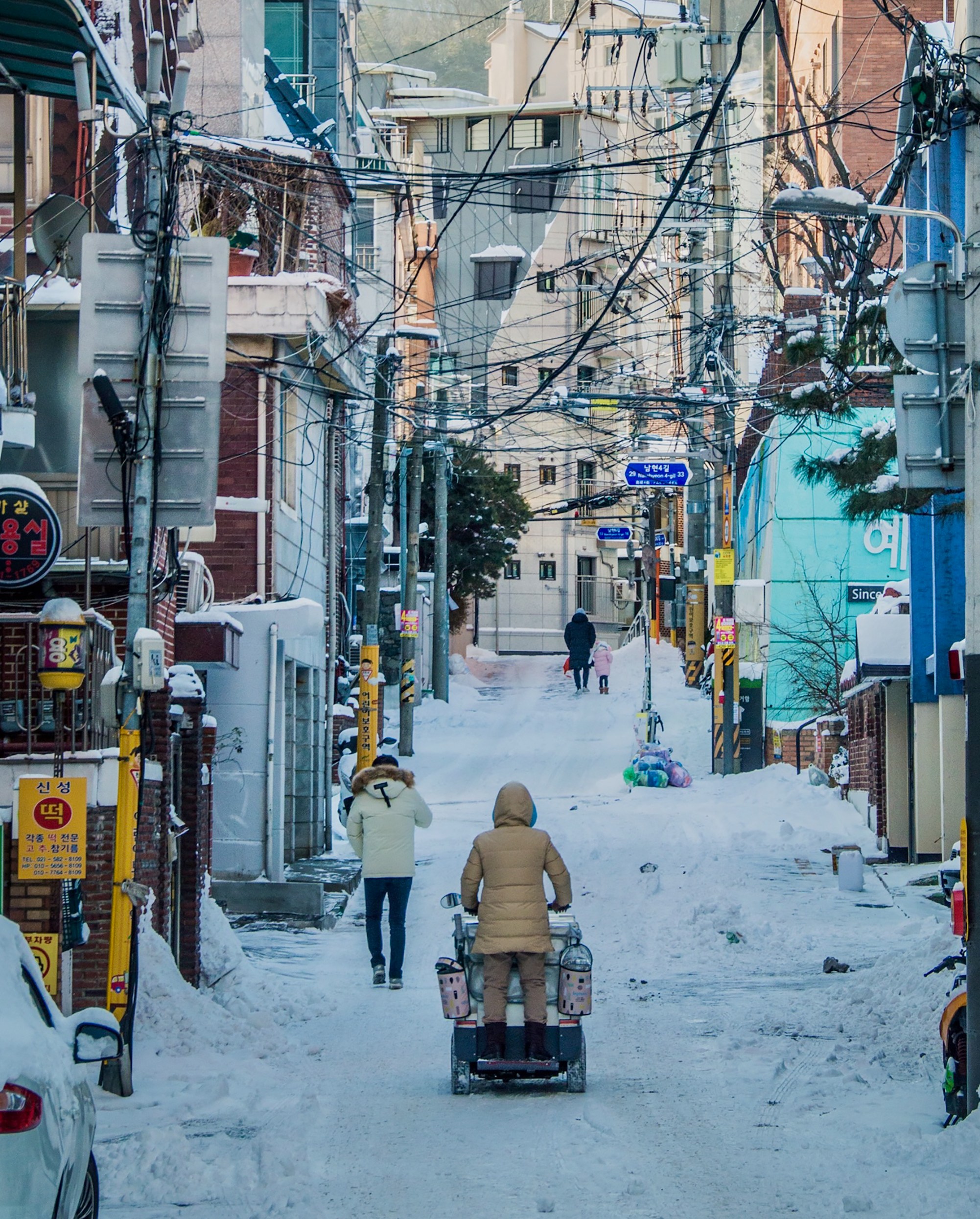 A woman in a thick coat rides a scooter up a snowy urban hill.