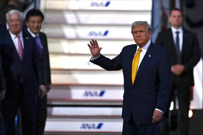 Person in formal attire waves while descending airplane steps, flanked by other individuals. Security personnel stand nearby