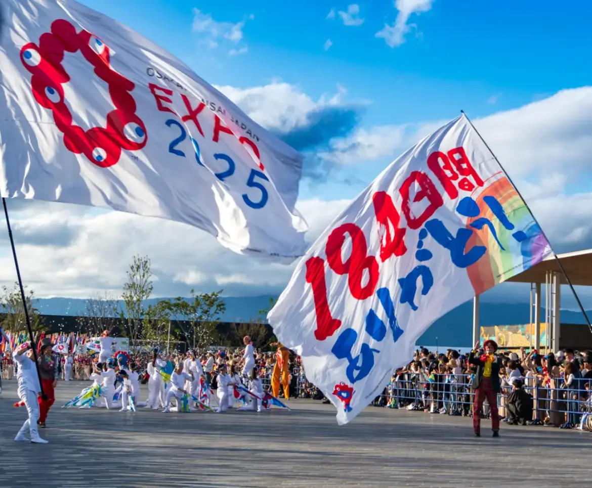 Performers waving large Expo 2025 Osaka flags during the closing celebration with crowds watching under a bright sky.