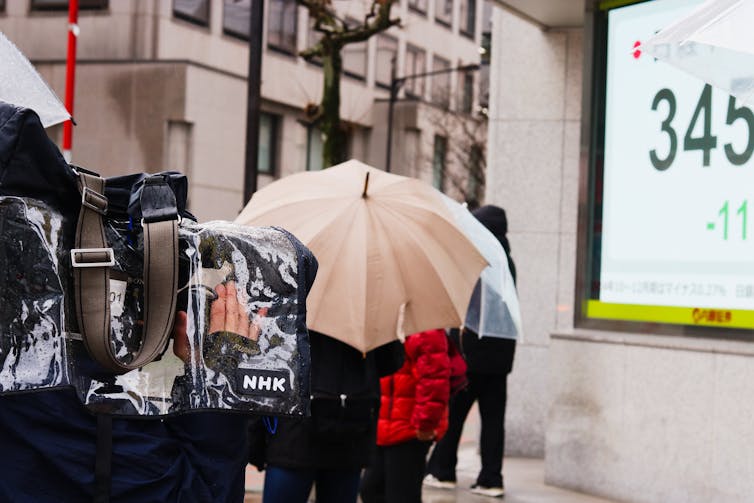 An NHK cameraman films a reporter with an umbrella on a rainy day