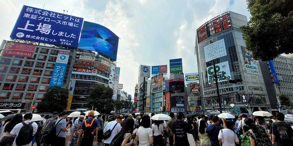 Busy urban intersection with a crowd crossing the street, surrounded by tall buildings covered in Japanese billboards and advertisements, including signs for IKEA and UC. Some pedestrians carry umbrellas under a partly cloudy sky.