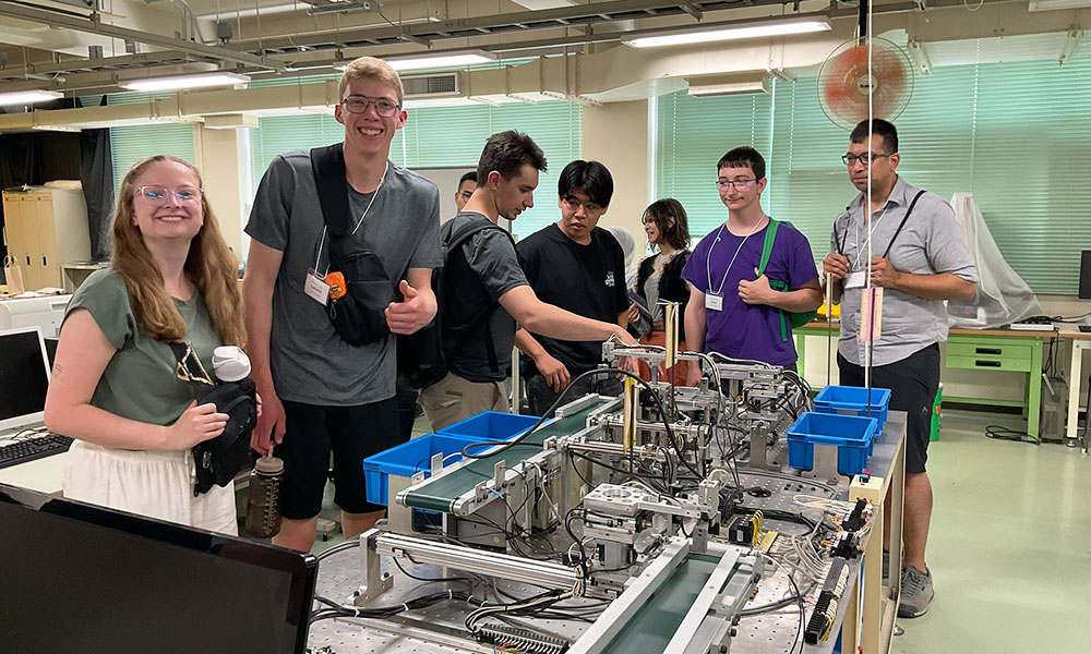 Group of people gathered around a mechanical setup in a workshop or lab, interacting with components including wires, blue containers, and machinery, suggesting a collaborative or educational activity.