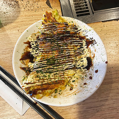 Plate of okonomiyaki, a Japanese savory pancake, topped with dark sauce, mayonnaise, and green herbs, placed on a wooden table beside chopsticks.