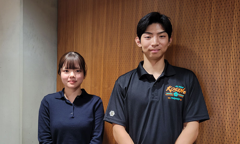 Two individuals standing side by side in front of a wooden panel wall. One wears a dark long-sleeve shirt, and the other wears a black polo shirt with colorful text and graphics including the word 'KOSEN.'