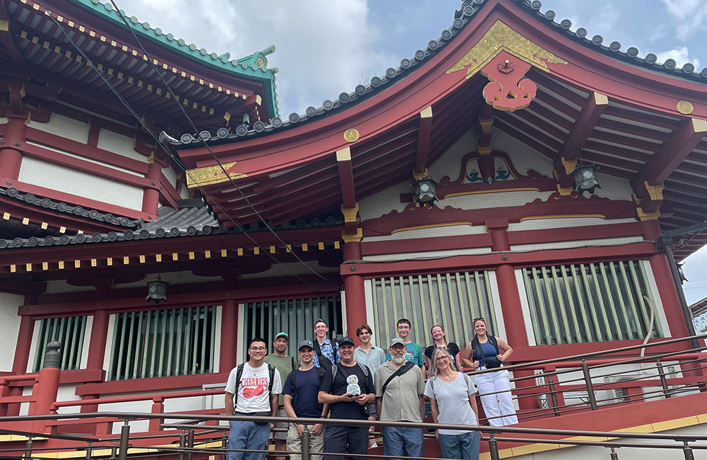 Group of people posing on a platform in front of a traditional Japanese building with red and white colors, ornate wooden beams, and a tiled roof.