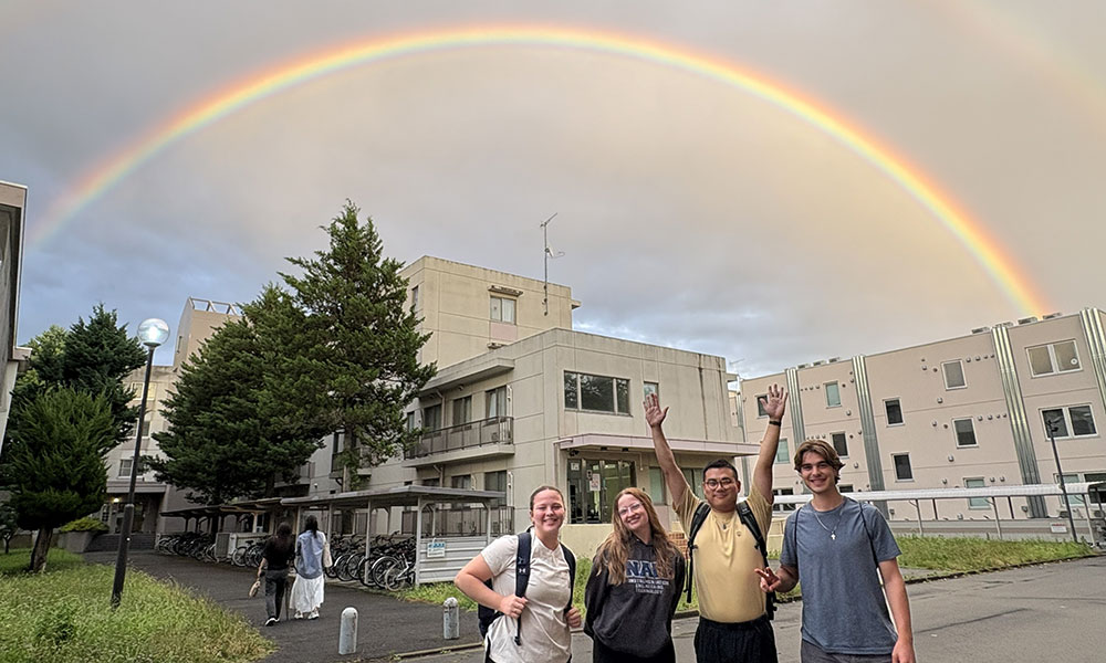 Four people standing outdoors in front of a building complex with a vivid rainbow arching across the sky. Trees, bicycles, and multiple buildings suggest a campus or residential setting.