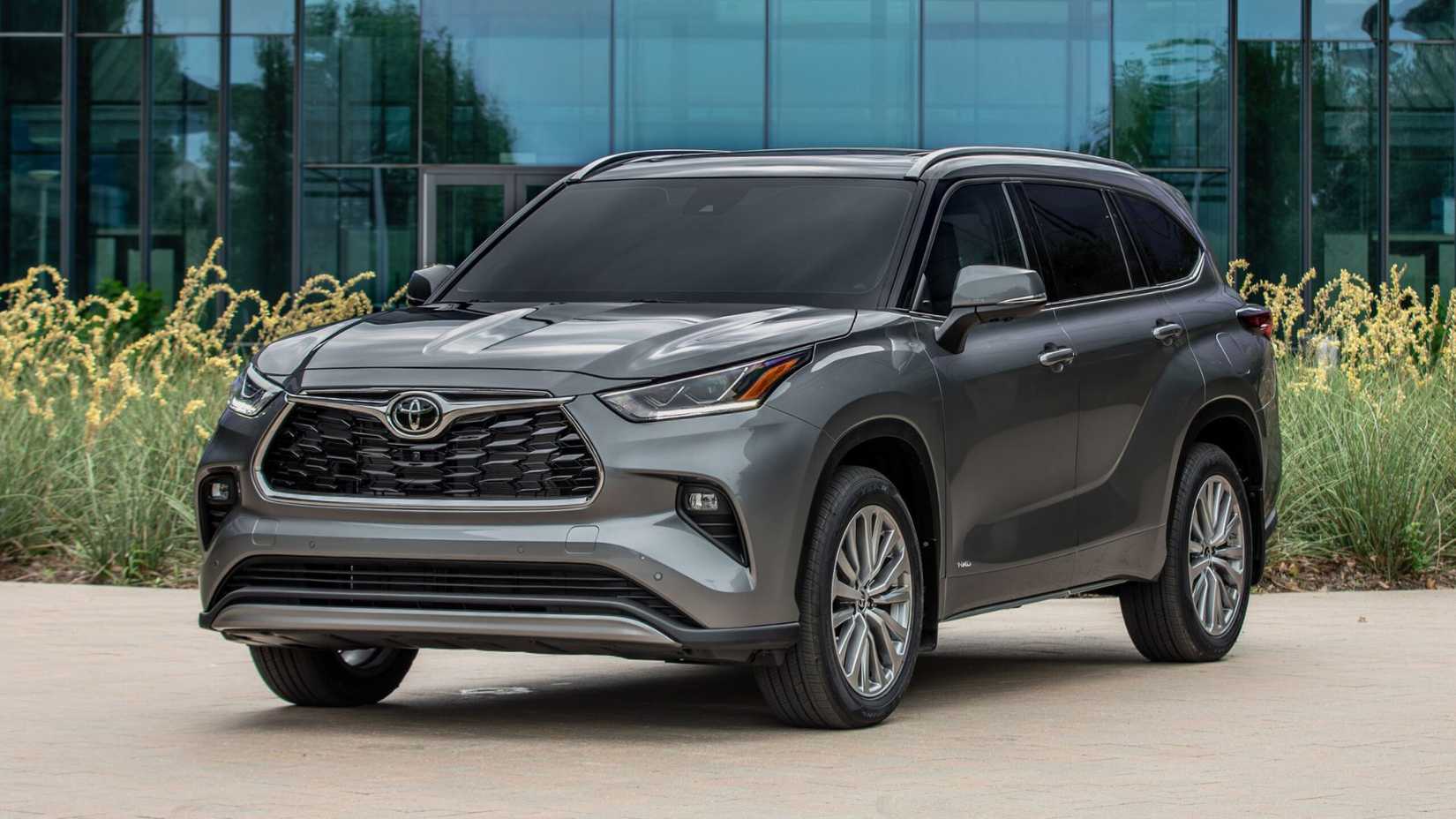 Static front 3/4 shot of a gray 2025 Toyota Highlander Hybrid parked in front of a modern glass-fronted building.