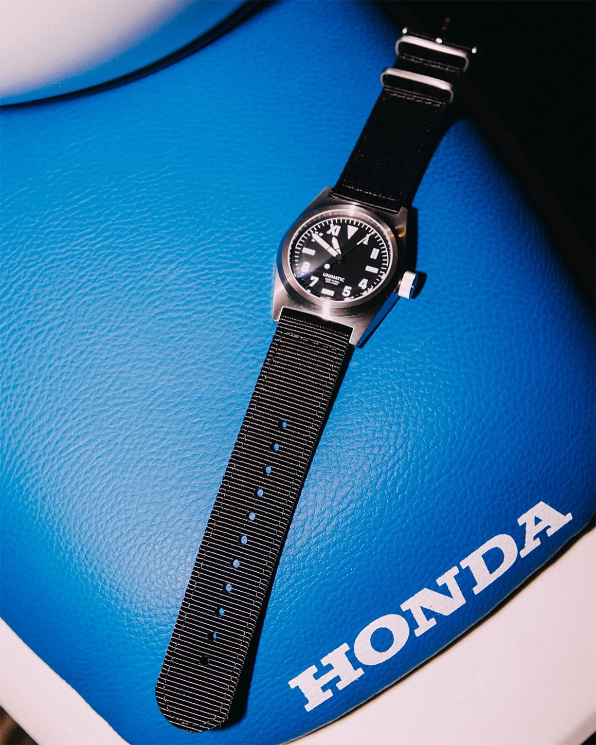 Black Unimatic wristwatch with a nylon strap placed on a blue leather surface with the word 