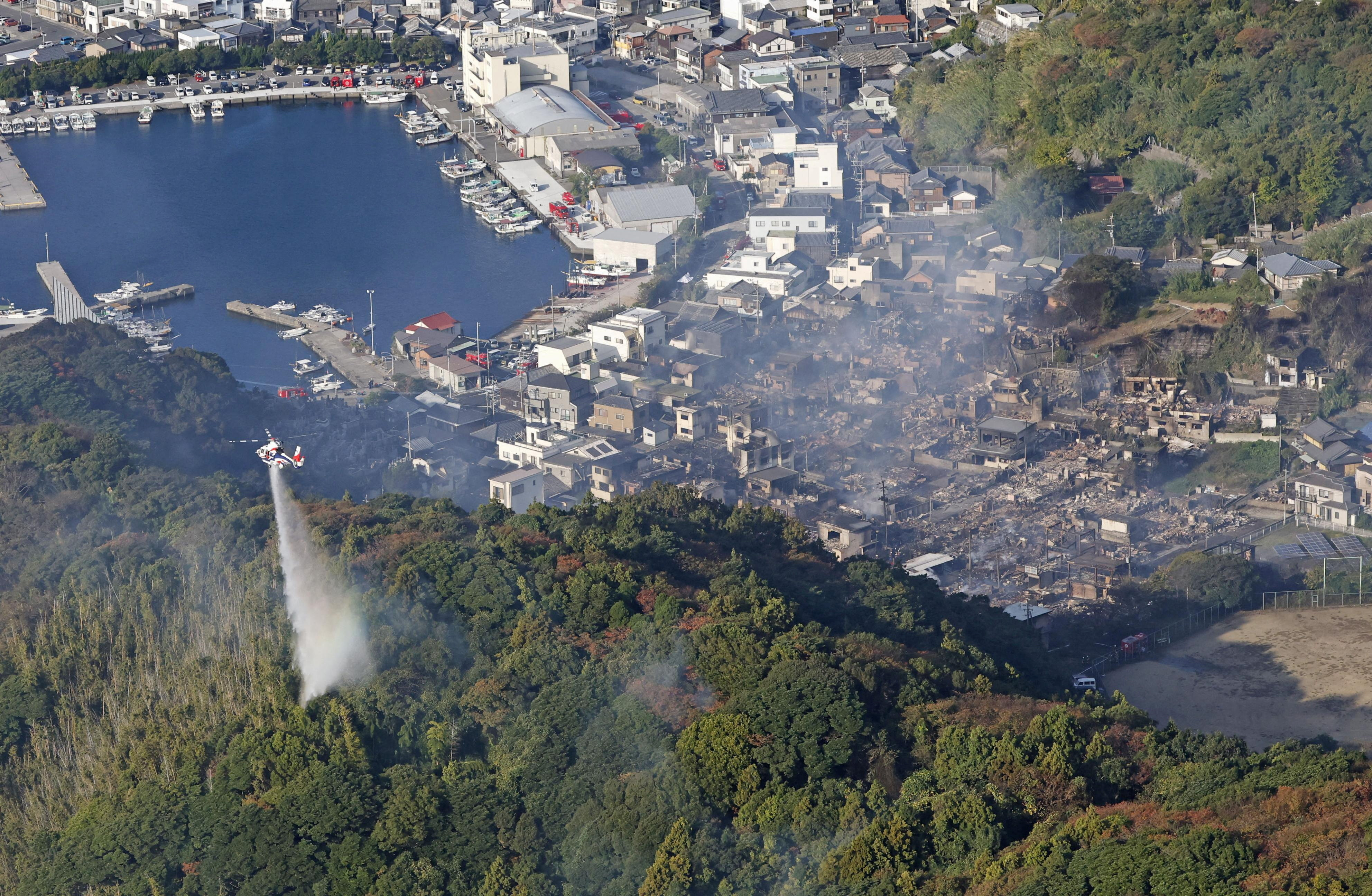 Aerial view of a helicopter dropping water on a forest fire with a destroyed town in the background and a harbor.