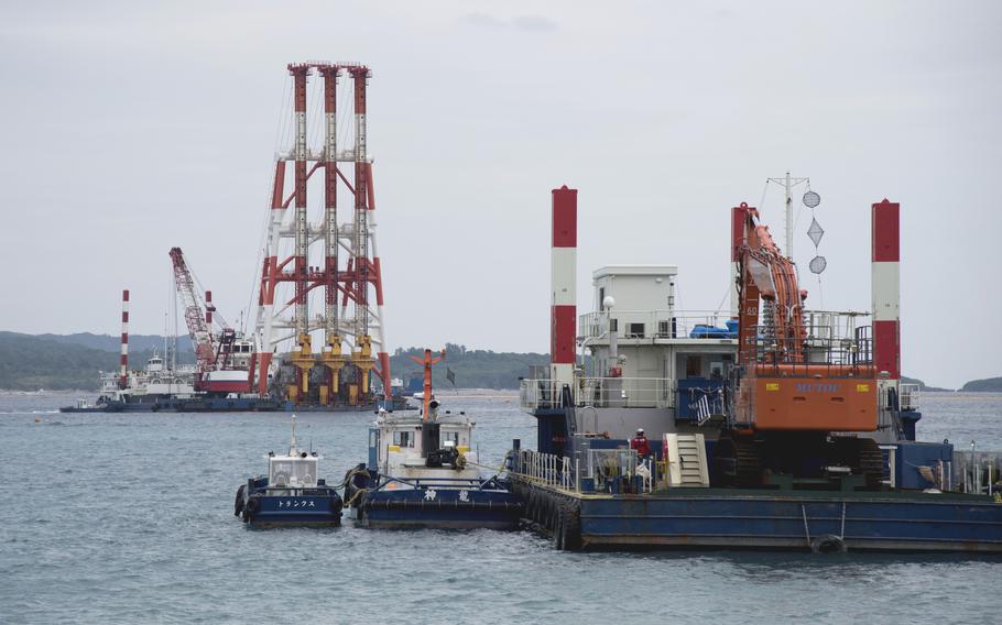 Two large maritime construction vessels sit in the water with smaller guide boats floating alongside them and a tree-lined shore in the background.
