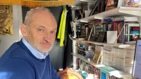 Nick Megoran is a man in his 40s or 50s with a receding hairline, grey hair and grey stubble. He is wearing a blue jumper and is sitting in a small study with lots of books on the shelves behind him. In the corner of the photograph you can see the edge of his computer 
