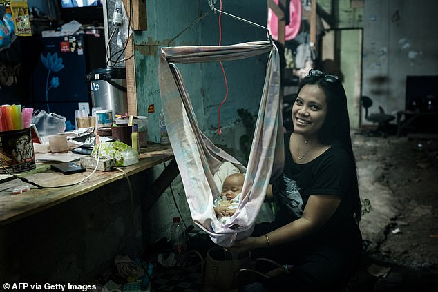 A mother poses as she soothes her two-month-old daughter placed in a baby bouncer next to a kiosk in Cilincing, North Jakarta, on November 5, 2025