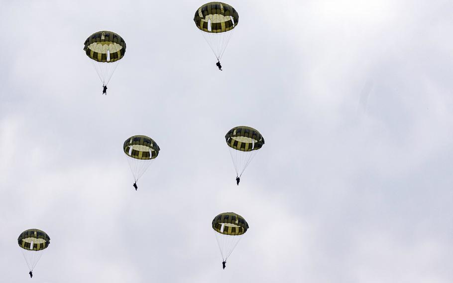 Six paratroopers, seen from below, descend toward the ground attached to olive green parachutes, with a cloud-filled skky in the background.