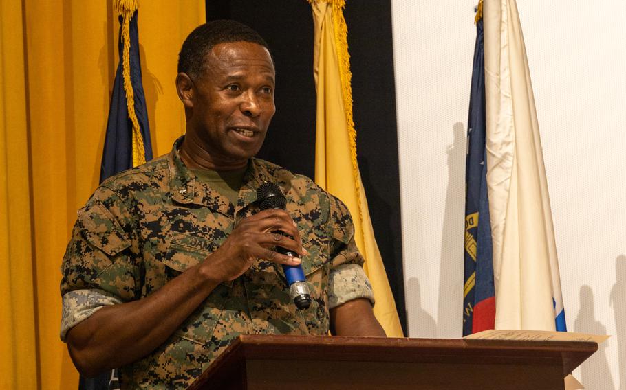 A Marine officer in camouflage uniform holds a microphone and speaks while standing at a podium in front of a yellow and black curtain backdrop.