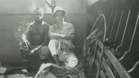 Trudy Johnson Two men, including Major Yutaka Umezawa, are sitting in the bottom of the boat. Both are smoking. Major Umezawa is in white wearing a white hat, shirt and trousers. The other man, an American soldier, is bare-chested. 