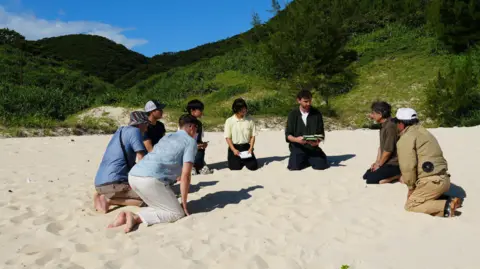 Nick Megoran A group of 8 people are kneeling in the sand on a beach on Aka island in Japan. Behind them is a steep cliff covered in green vegetation 