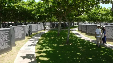 Nick Megoran The photograph shows the Cornerstone of Peace Memorial in Japan which commemorates those lost during the Battle of Okinawa. Large granite blocks are arranged in rows in concentric rings and on each are dozens of names. A Japanese couple walk along the path gazing at the names. Many trees give shelter.