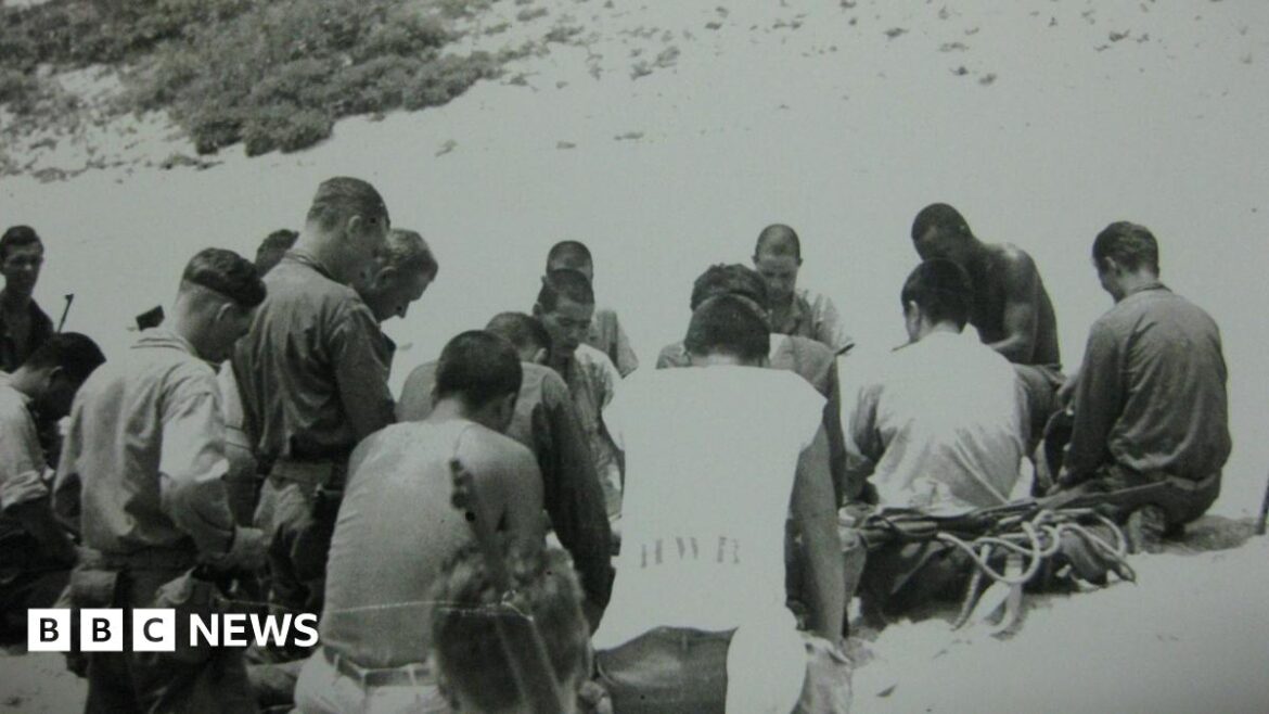 Newcastle professor recalls US-Japan picnic truce A black and white photograph shows a group of about 20 men sitting and kneeling in prayer on a beach. Some of the men are Japanese, some are American. They all have short hair and have their heads bowed.