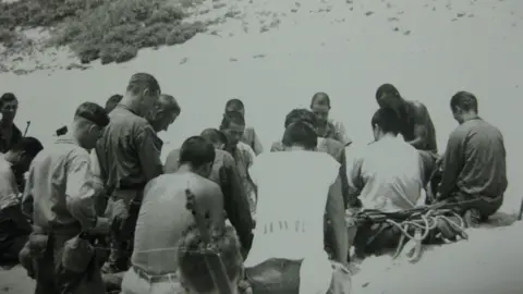 Trudy Johnson A black and white photograph shows a group of about 20 men sitting and kneeling in prayer on a beach. Some of the men are Japanese, some are American. They all have short hair and have their heads bowed.
